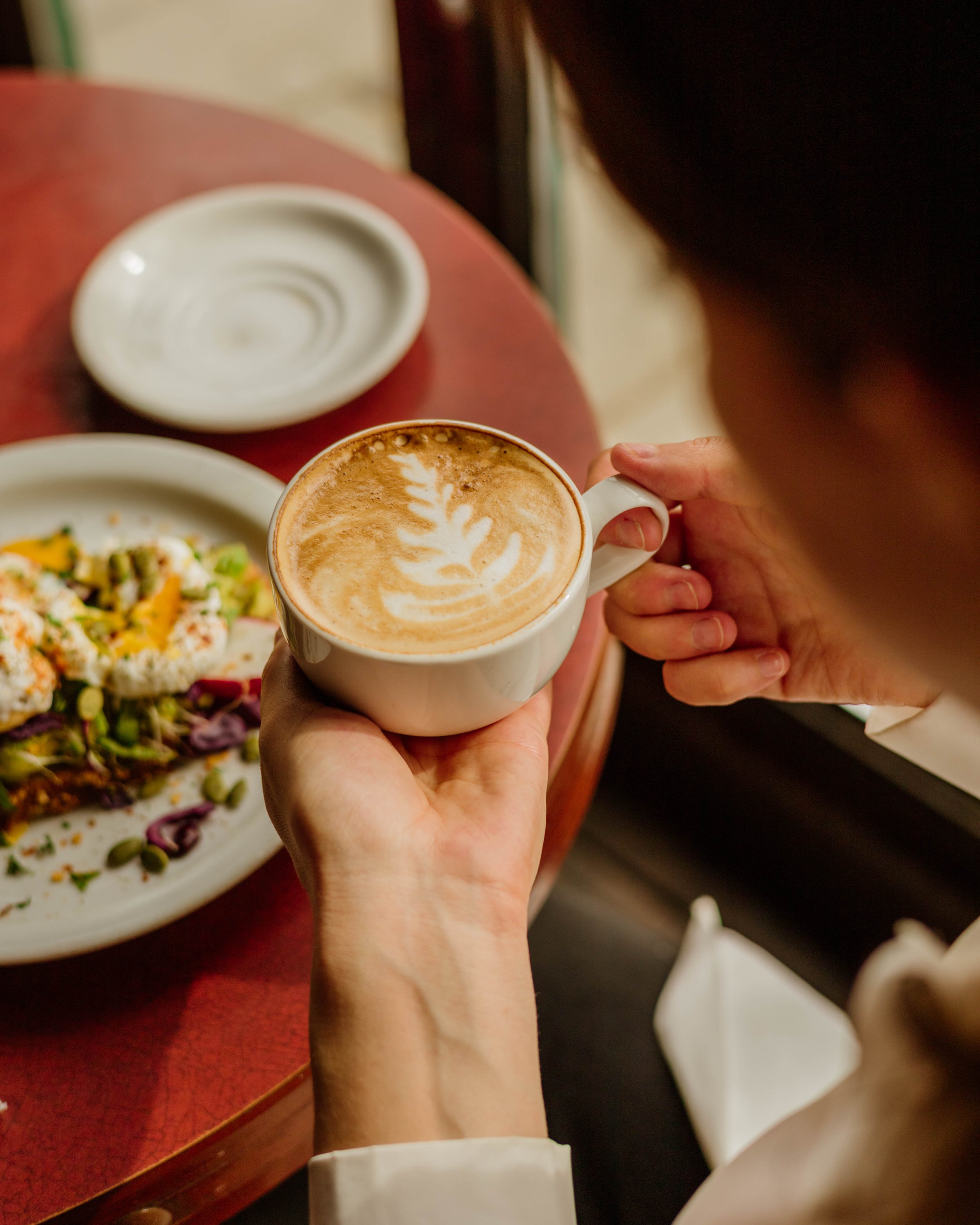 Une personne tient une tasse de cappuccino avec un motif de feuille dans un café, avec une tartine à l'avocat et oeuf poché